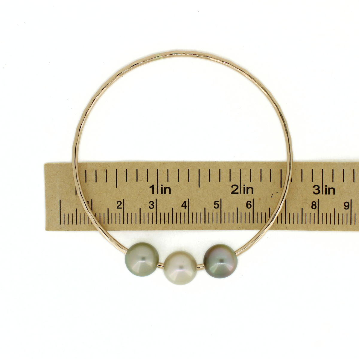 A gold bangle bracelet with three round Tahitian pearls in shades of gray-green, displayed on a paper ruler against a white background.
