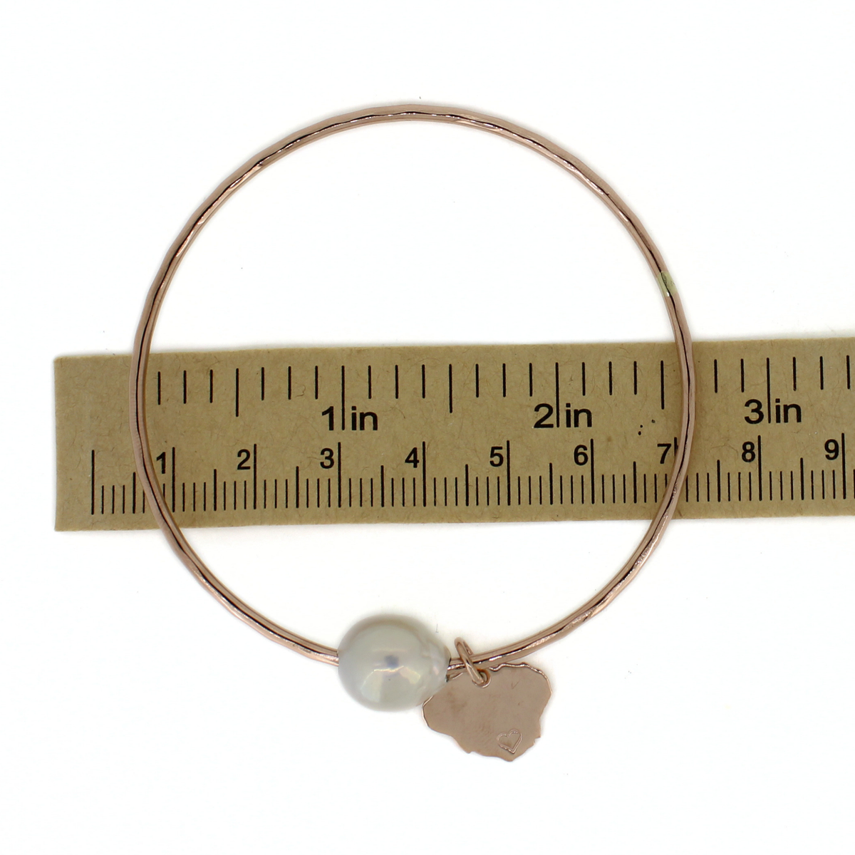 A thin rose gold wire bangle bracelet with a white pearl bead and a small hand-forged rose gold Kauai charm, shown next to a paper ruler for scale.