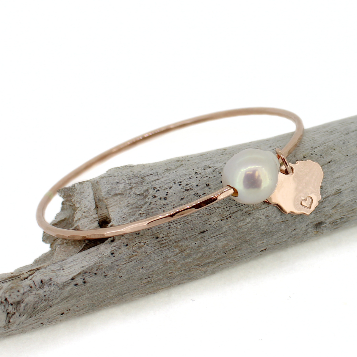 A delicate rose gold bangle bracelet with a white freshwater pearl and a small rose gold Kauai charm, displayed on a piece of weathered driftwood against a white background.