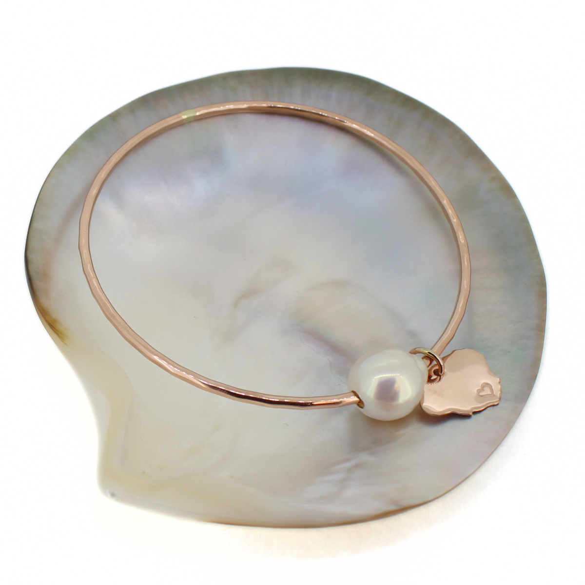 A rose gold bangle bracelet with a white pearl and a small Kauai island charm, displayed on a white mother-of-pearl shell.