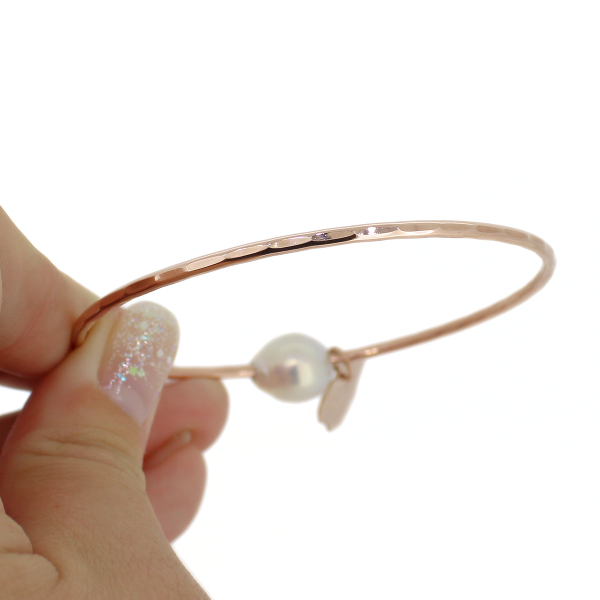 A close-up of a hand holding a thin rose gold bangle bracelet with a hammered texture, against a plain white background.