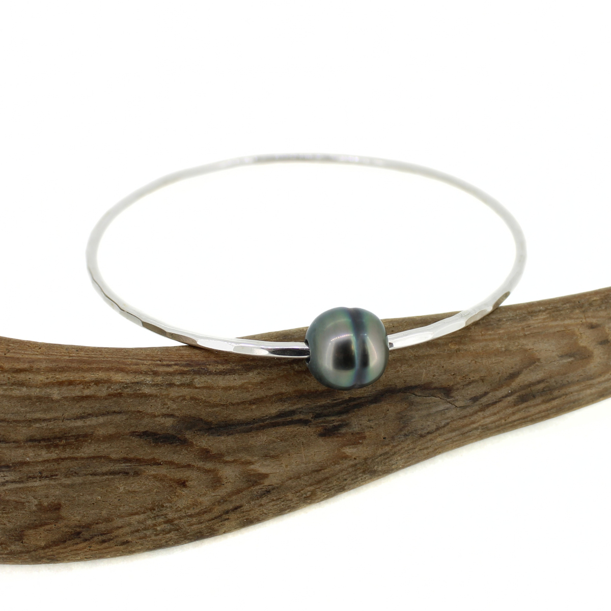 Round silver bangle with a dark Tahitian pearl displayed on a piece of driftwood