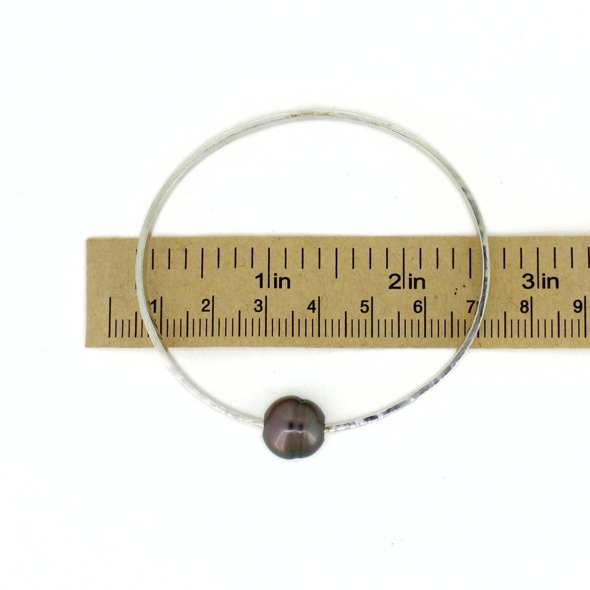 A silver oval bangle bracelet with a single dark gray pearl, shown horizontally over a wooden ruler for scale on a white background.
