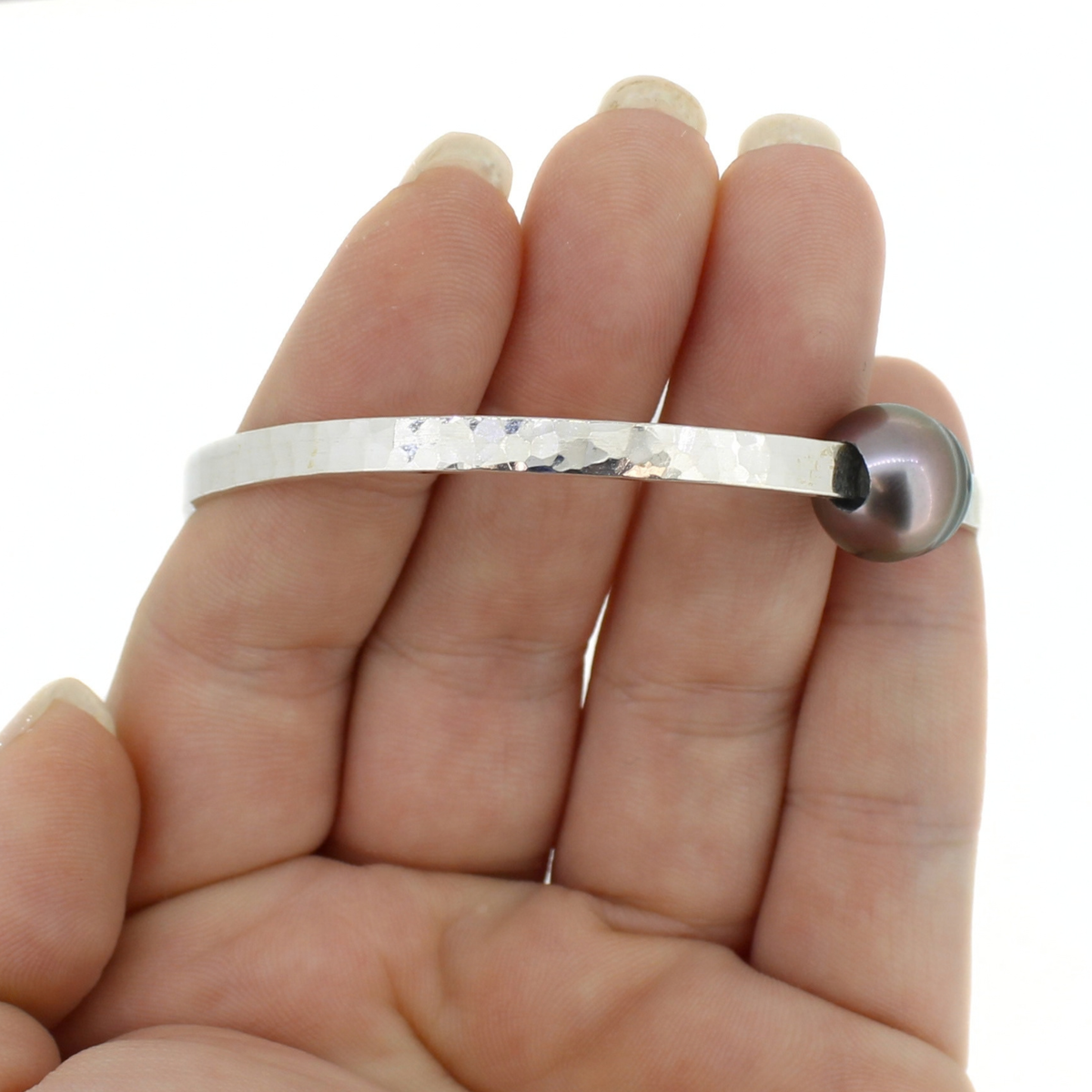A silver hammered metal bangle bracelet with a dark gray round pearl, shown resting on a person's hand against a white background.
