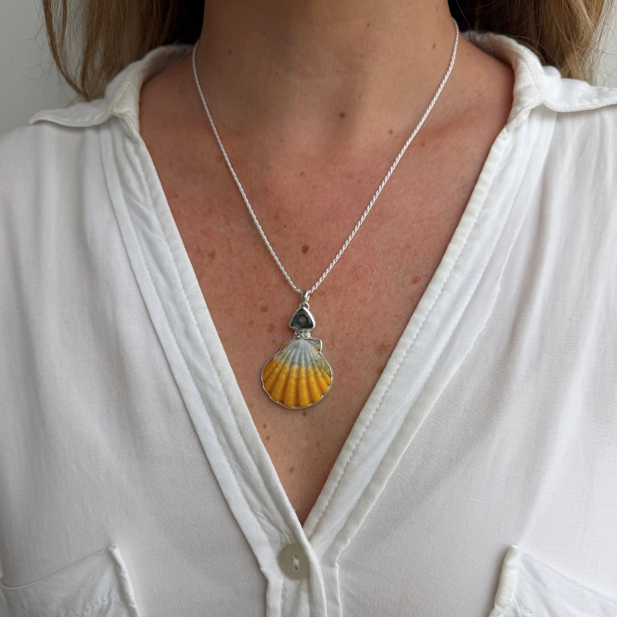 A close-up of a person wearing a silver chain necklace with a sunrise shell pendant featuring blue tourmaline gemstone above it, styled with a white button-down shirt.