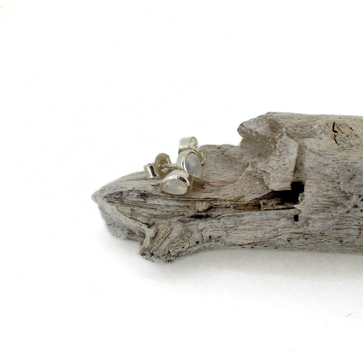 A pair of small silver stud earrings with oval clear stones, resting on a piece of weathered driftwood against a plain white background.