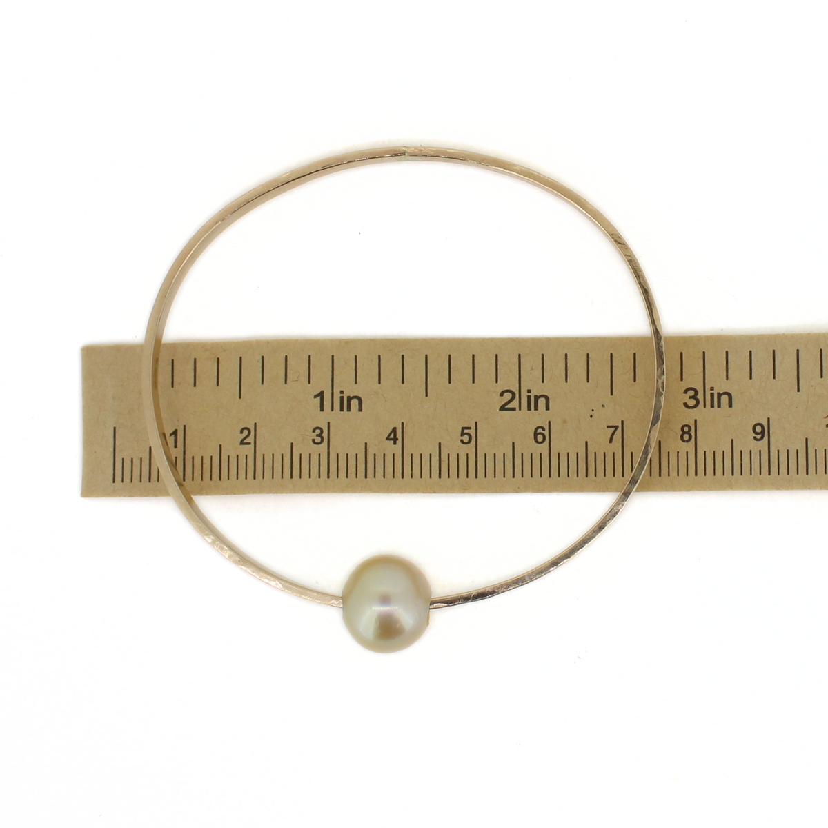A thick gold bangle bracelet with a single gold pearl bead, shown next to a ruler for scale on a white background.