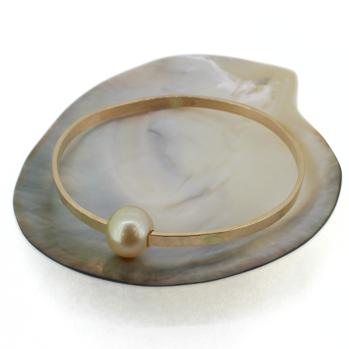 A thick gold bangle bracelet with a single round pearl, displayed on a mother-of-pearl shell tray against a white background.