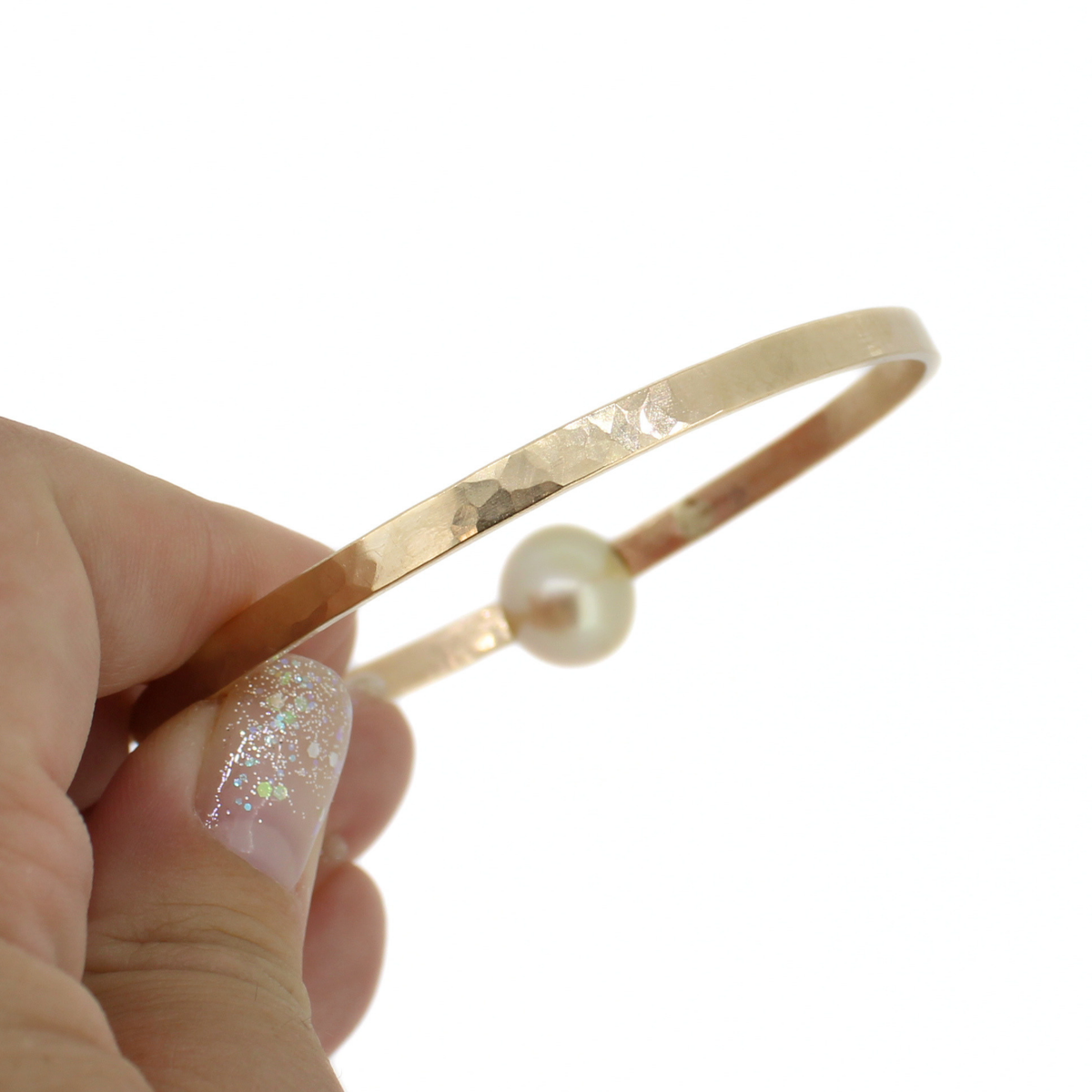 A hand with glittery nail polish holds a thin hammered gold-tone bangle bracelet with a single gold pearl bead against a plain white background.