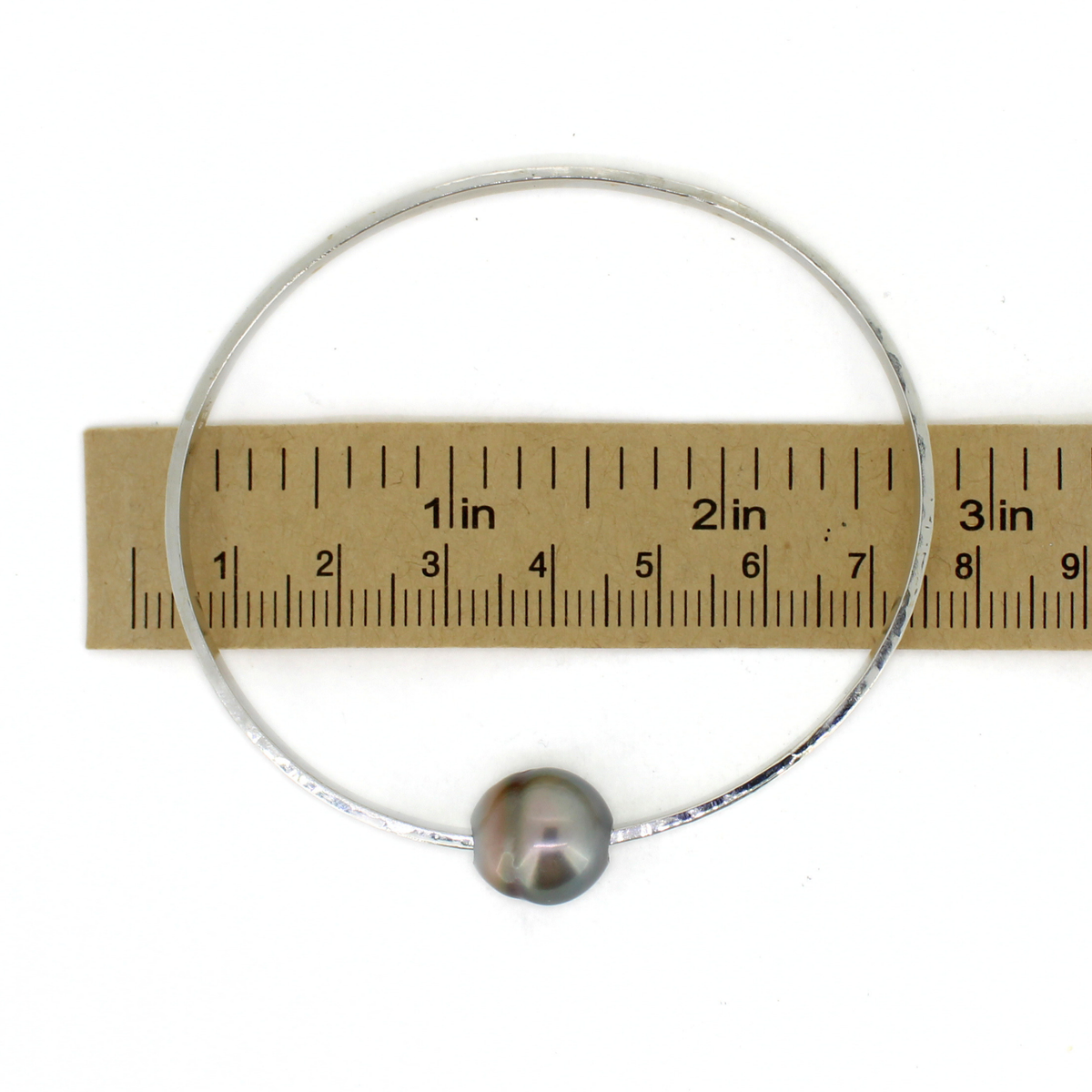A thick silver bangle bracelet with a single gray pearl, shown next to a brown paper ruler for scale on a white background.