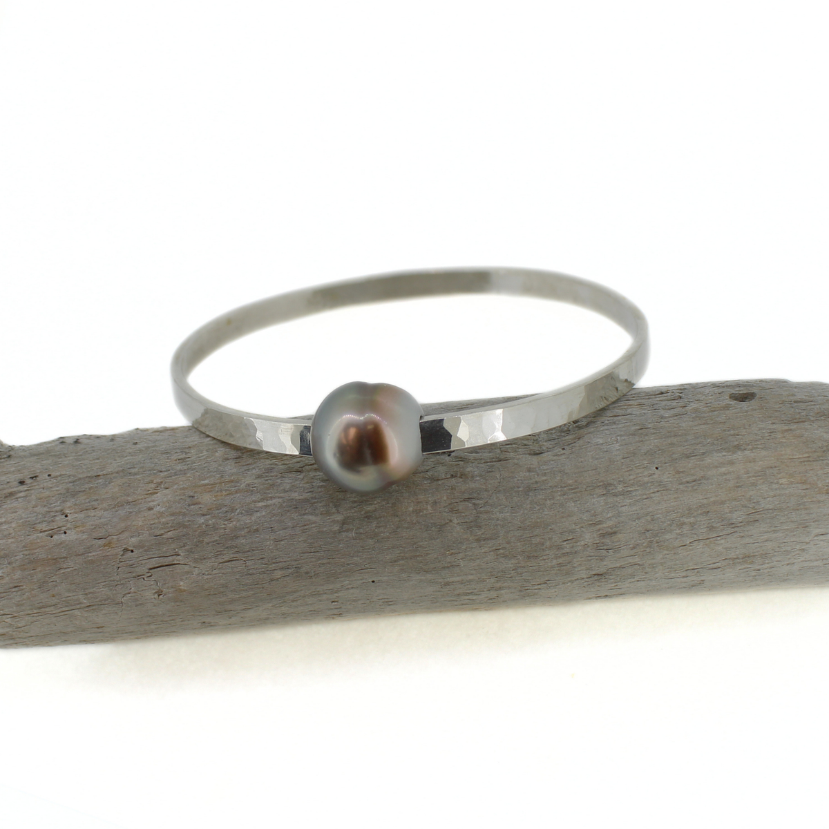 A sterling silver bangle bracelet with a grey Tahitian pearl displayed on a piece of driftwood agains a white background