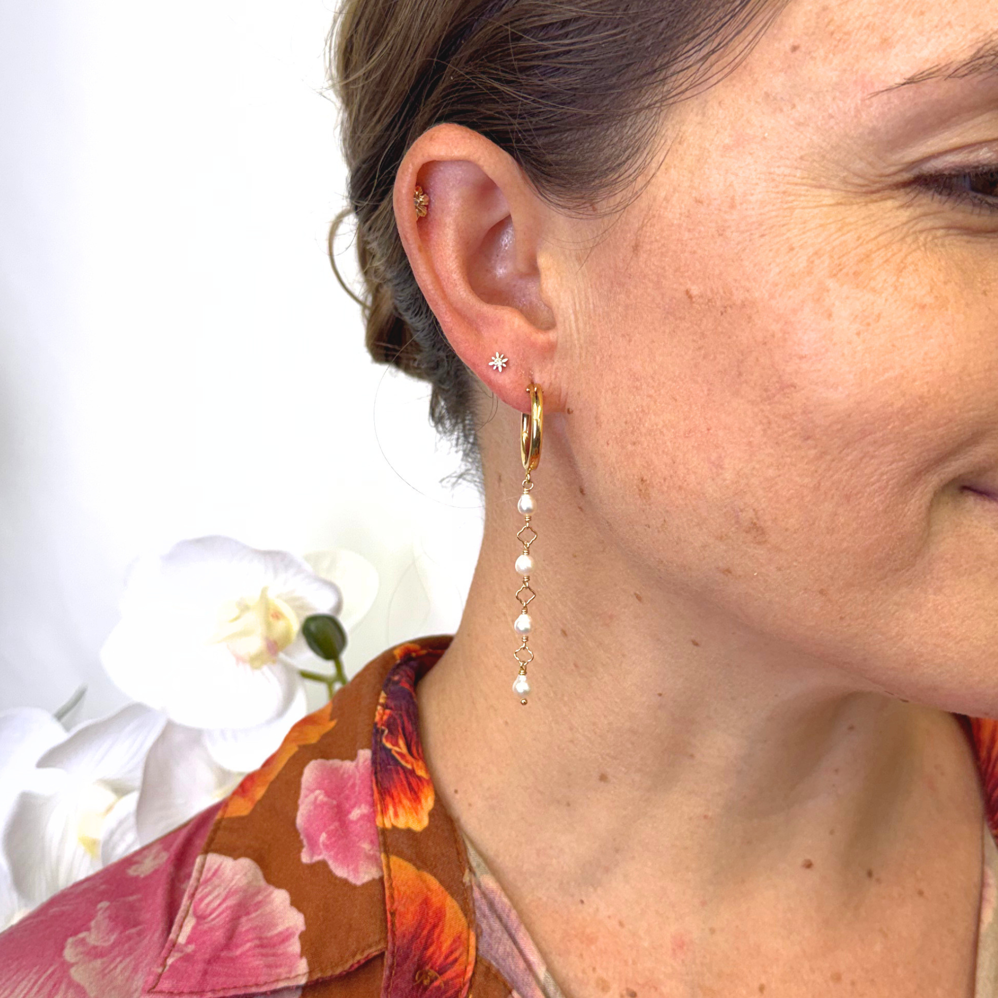 A close-up of a woman's ear wearing a delicate gold huggie hoop earring with a dangling chain of small white pearls and a tiny gold star stud.