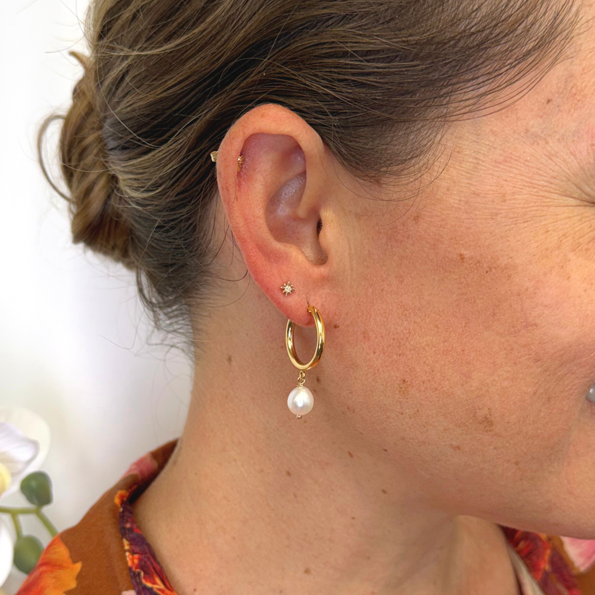 A close-up of a woman's ear showcasing a gold hoop earring with a dangling white pearl, along with other small ear piercings.