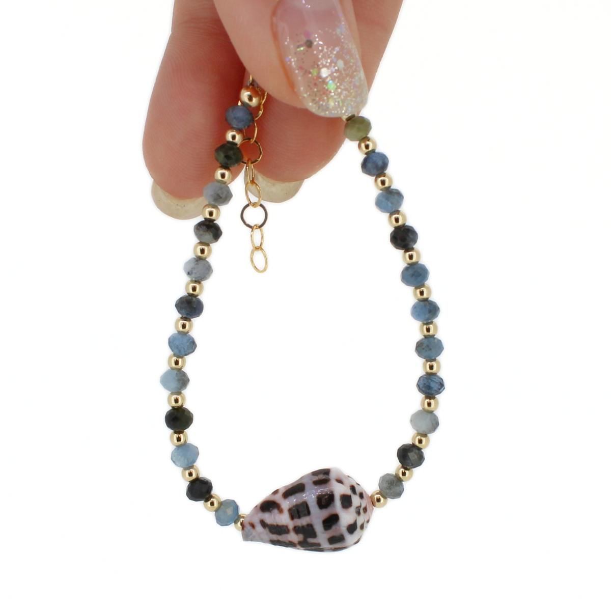 A delicate beaded bracelet with light and dark blue faceted beads, small gold spacer beads, and a large patterned seashell at the center, held by a hand against a white background.