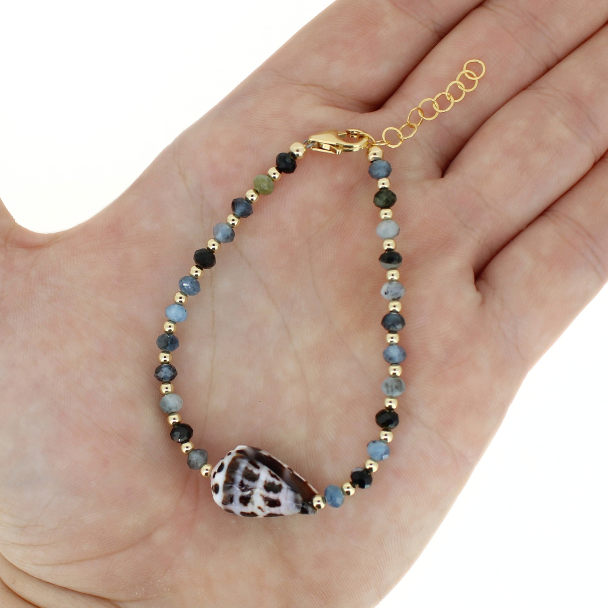 A beaded bracelet with a seashell at the center displayed on an open hand. The bracelet features faceted beads in light and dark blue, accented with small gold spacer beads and a gold clasp, with an adjustable gold chain extender.