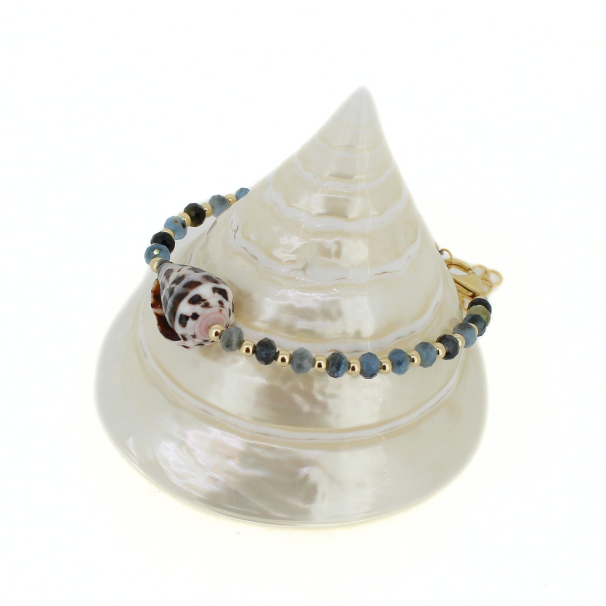 A beaded bracelet with blue-gray faceted beads, small gold spacer beads,  and a larger pink-and-black patterned shell charm, displayed on a white seashell stand against a white background.