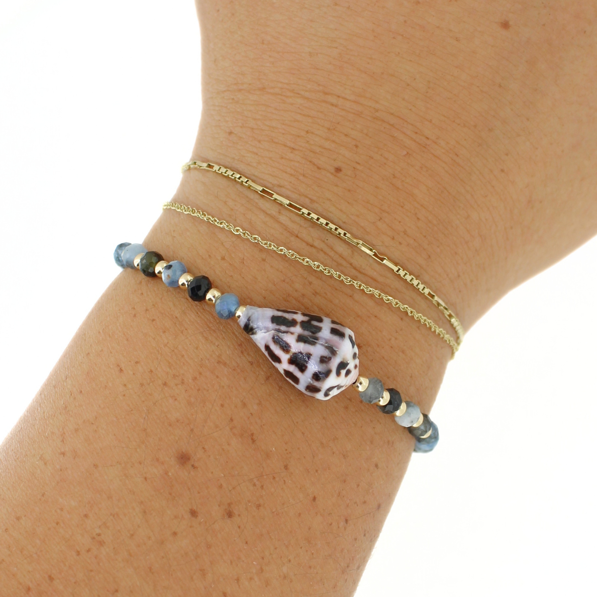 A close-up of a person's wrist wearing two delicate bracelets: a thin gold chain and a beaded bracelet featuring a large teardrop-shaped white shell with black spots, flanked by small blue and gold beads.
