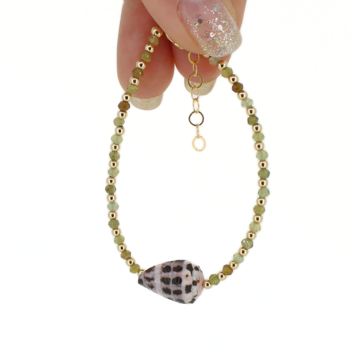A delicate beaded bracelet with light green and olive beads, gold spacer beads, and a white and black seashell at the center, held by a hand against a white background.