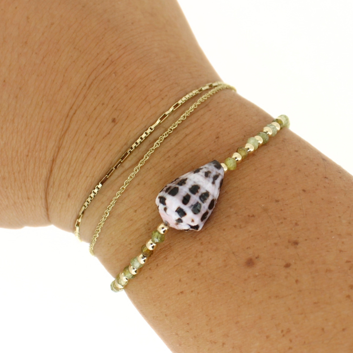 A close-up of a person's wrist wearing two delicate bracelets: one gold chain bracelet and one beaded bracelet featuring a white teardrop shell with black spots and small gold and green beads.