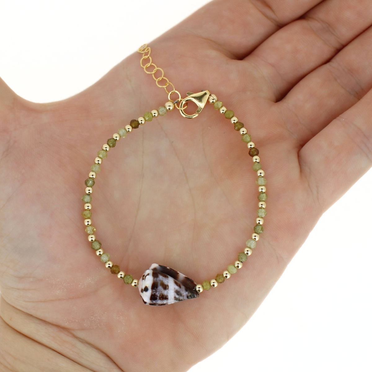 A delicate beaded bracelet with olive green and gold beads, a gold lobster clasp, and a brown and white patterned seashell, displayed on a person's palm.