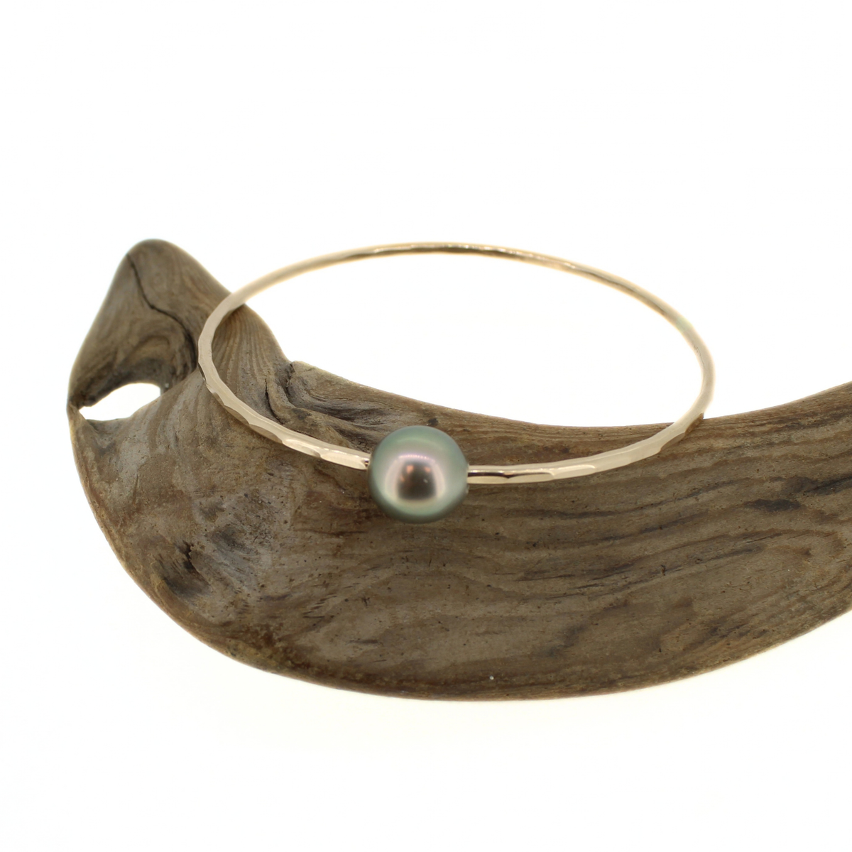 Gold hammered bangle with a green Tahitian pearl, displayed on a piece of driftwood