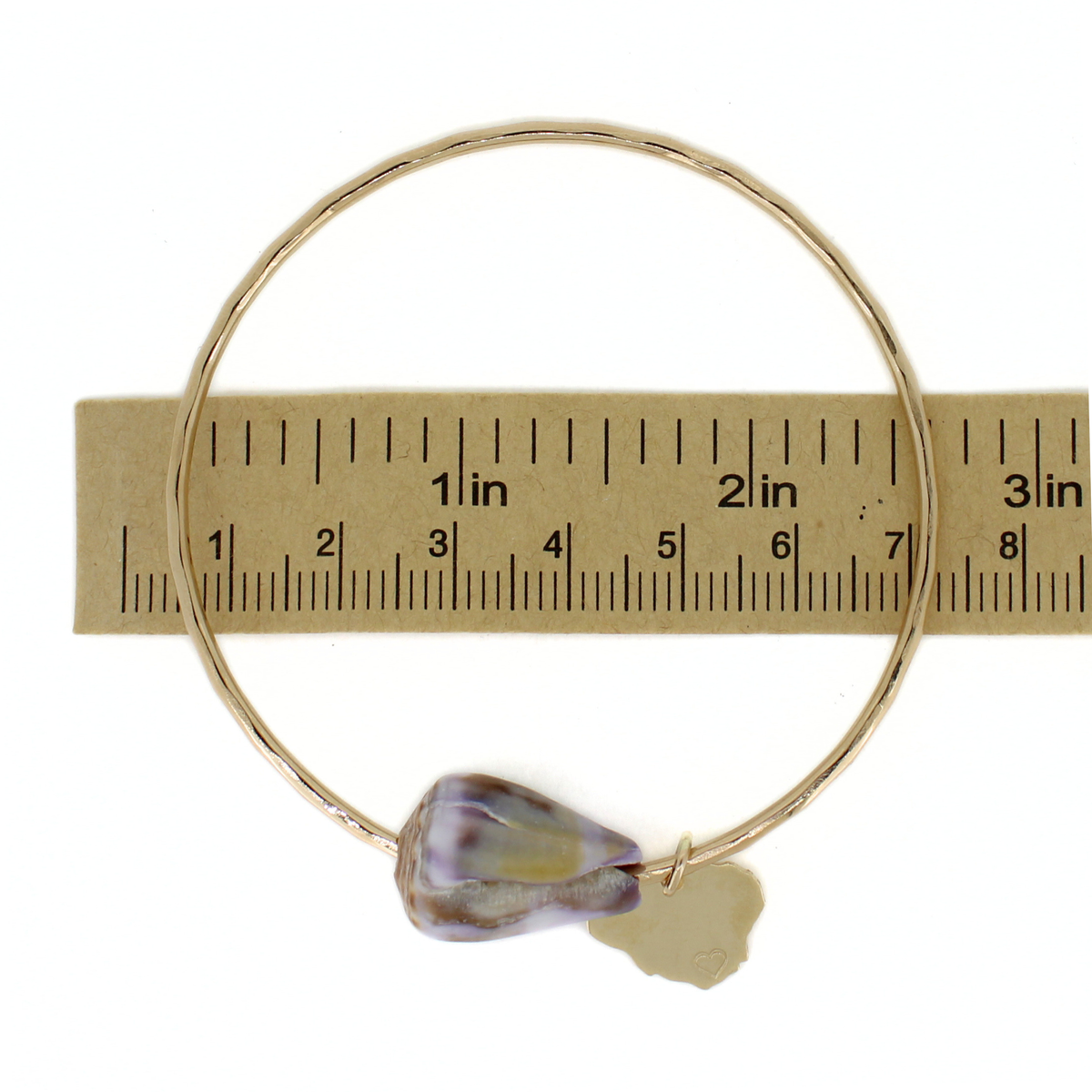 A thin gold bangle bracelet with a cone shell and Kauai charm, shown next to a brown paper ruler for scale on a white background.