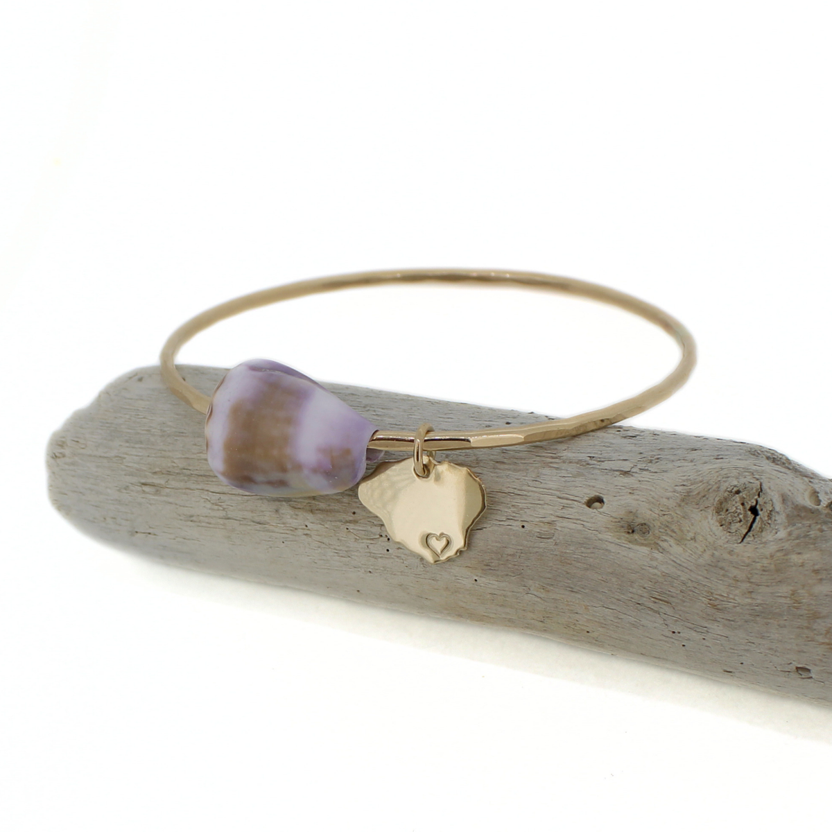 A gold bangle bracelet with a lavender and brown cone shell and a small gold Kauai-shaped charm, displayed on a piece of driftwood against a white background.