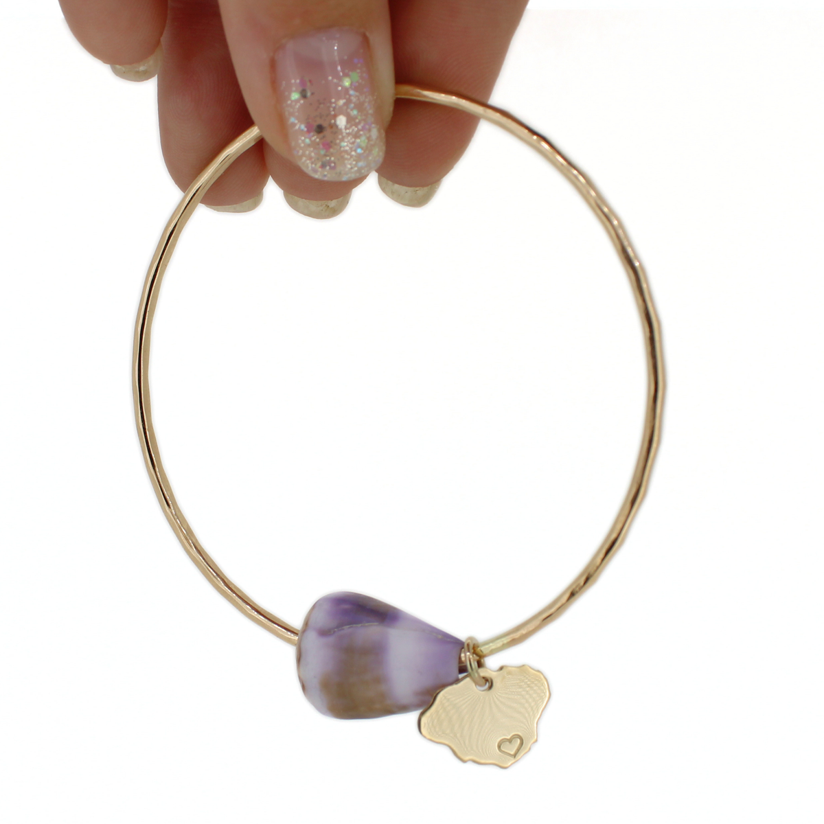 A gold bangle bracelet with a purple and white cone shell and a small gold Kauai charm, held by a hand with glittery nail polish against a white background.