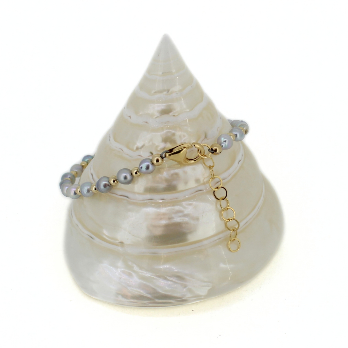 A beaded bracelet with gray pearls and gold spacer beads, featuring a gold clasp and extender chain, displayed on a white seashell stand against a white background.