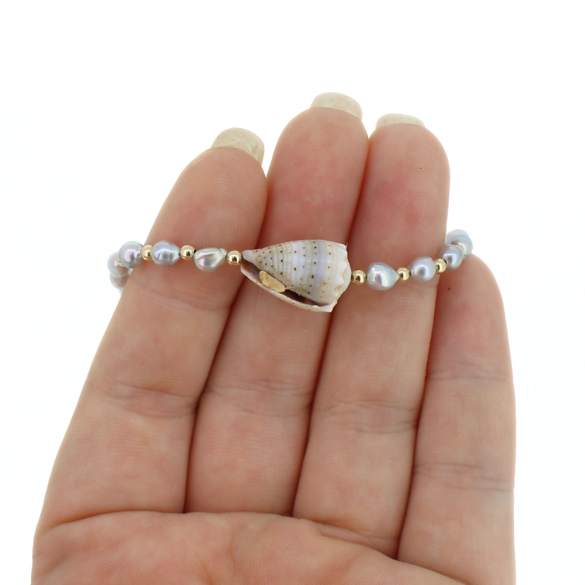 A beaded bracelet featuring a central seashell and small pearl-like beads with gold spacer accents, shown worn on a person's hand against a white background.
