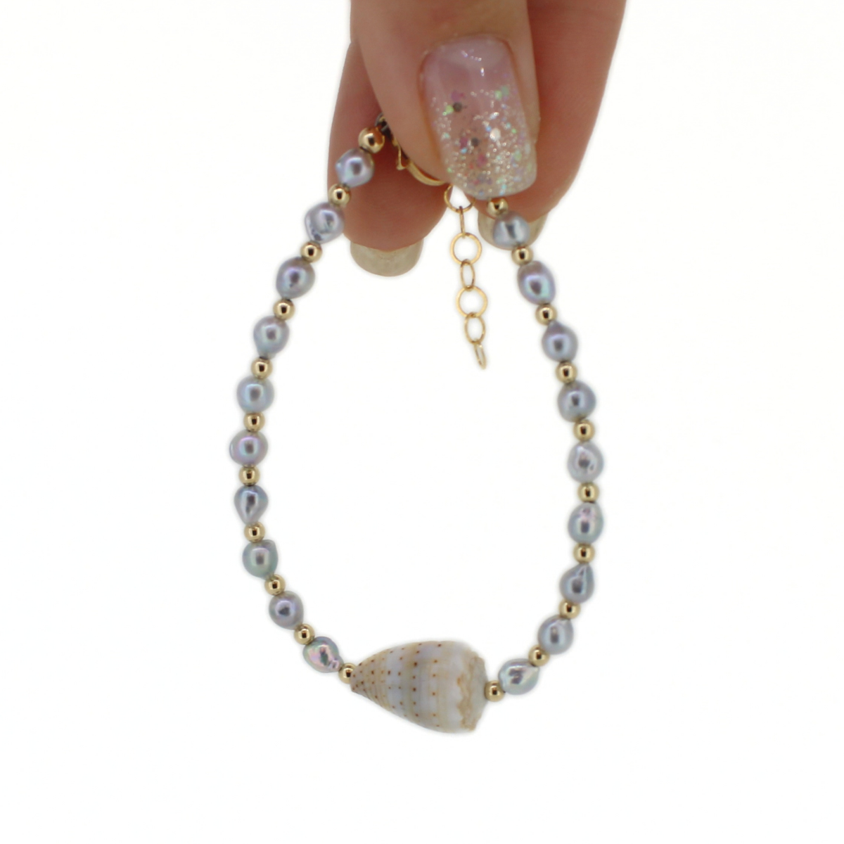 A delicate beaded bracelet with light gray pearls, small gold spacer beads, a gold clasp with extender chain, and a white seashell, held by a hand with glittery nail polish against a white background.