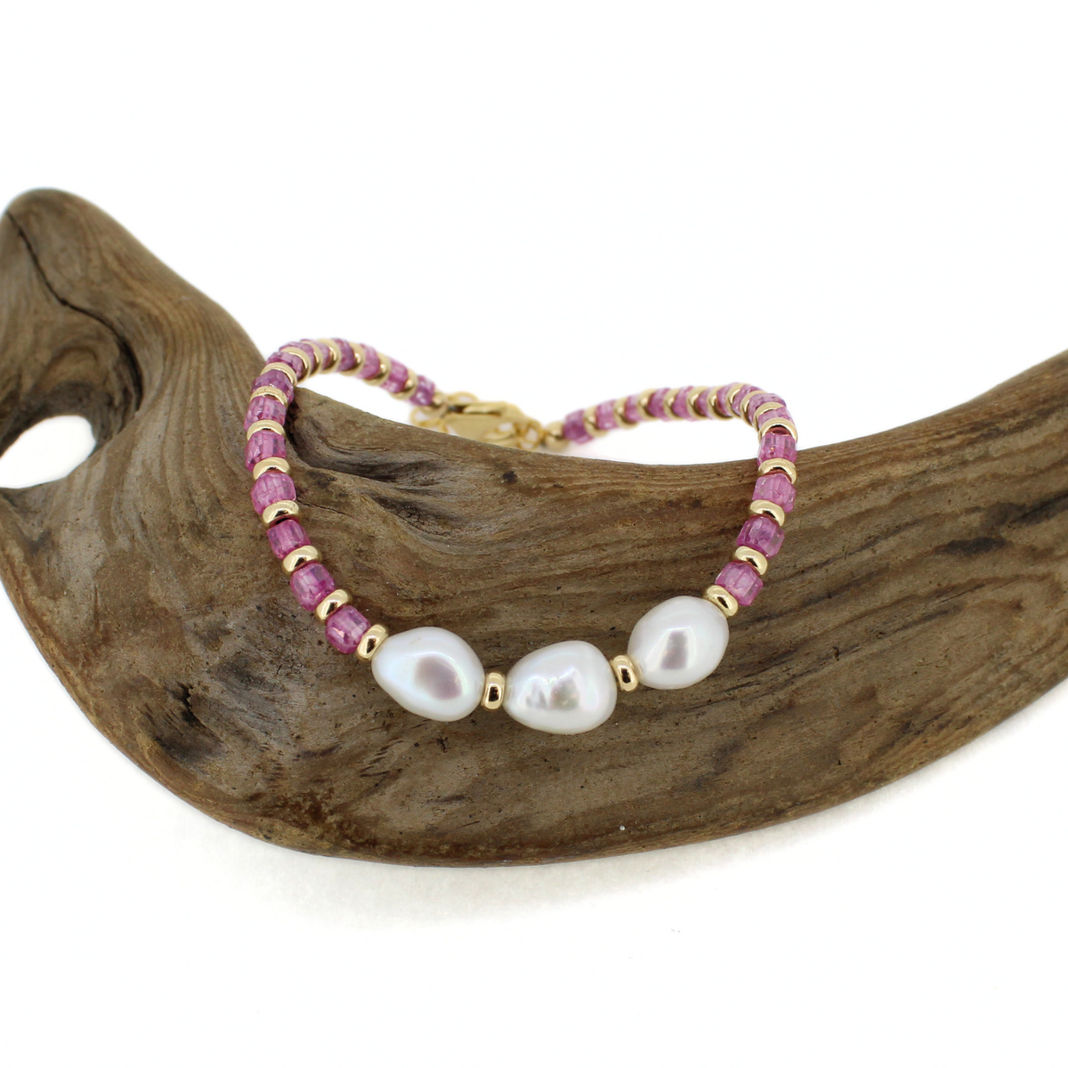 A beaded bracelet with three large white baroque pearls, pink gemstone beads, and gold spacer beads, displayed on a curved piece of natural wood against a white background.