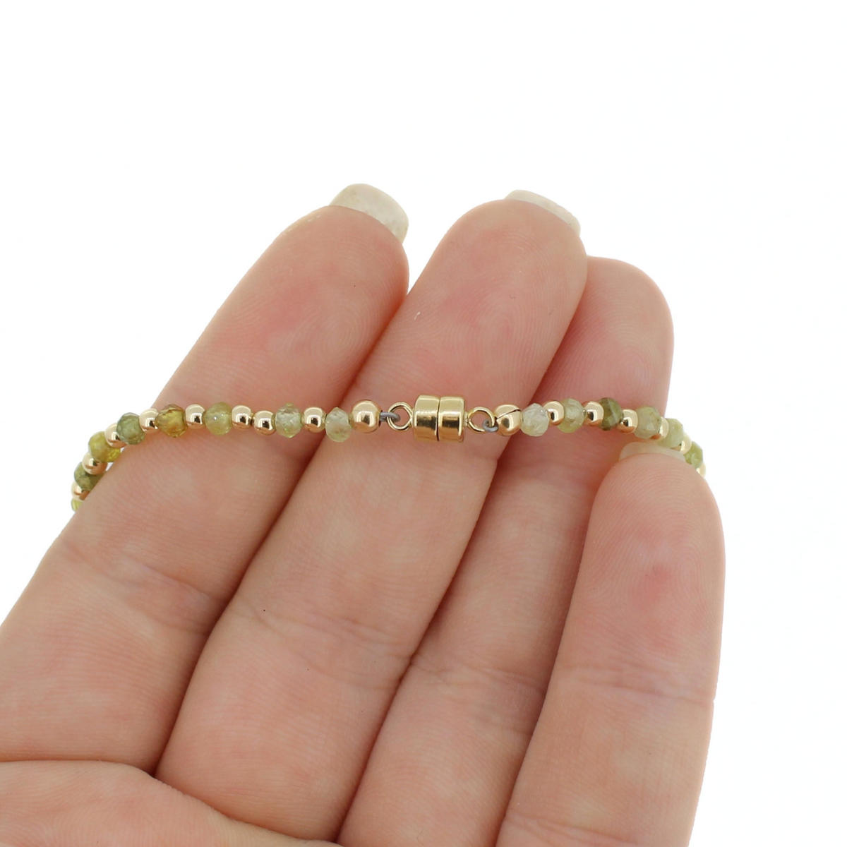 A delicate beaded bracelet with small gold and pale green beads, held on a person's fingers against a plain white background.