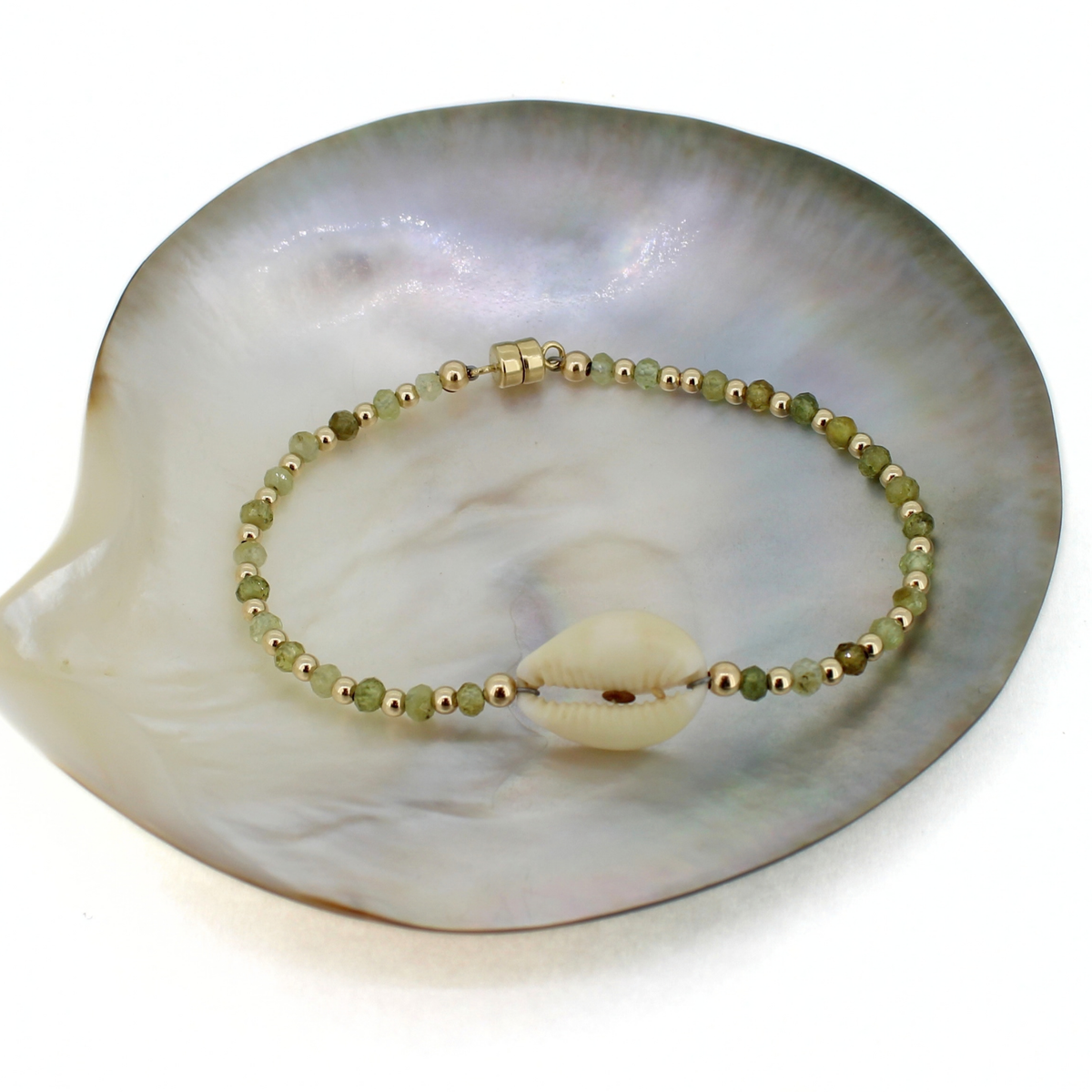 A beaded bracelet with small green and gold beads and a central cowrie shell, displayed on a large iridescent shell fragment against a white background.