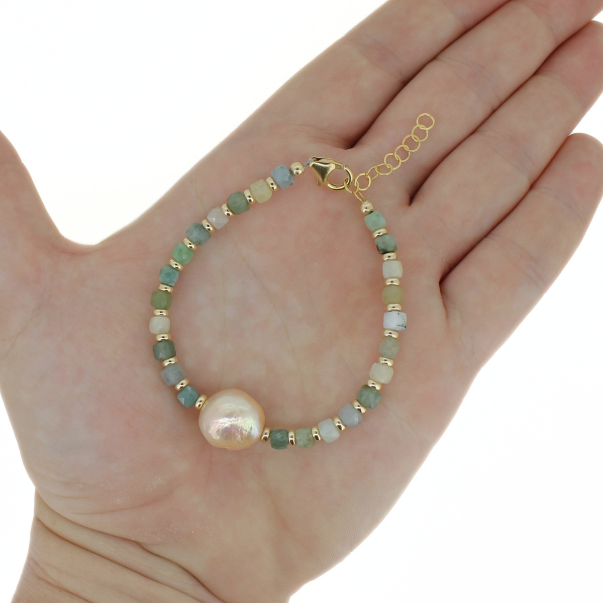 A gold-tone beaded bracelet with a large baroque pearl and small green and white beads, displayed on a person's open hand against a white background.