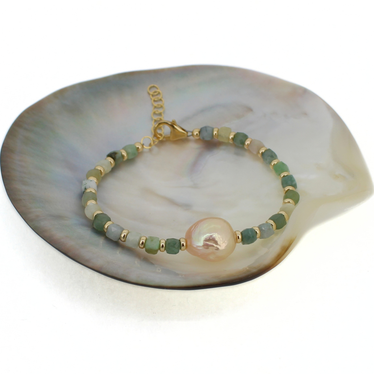 A beaded bracelet with green and gold beads, featuring a large iridescent cream colored pearl, displayed on a mother-of-pearl shell dish against a white background.