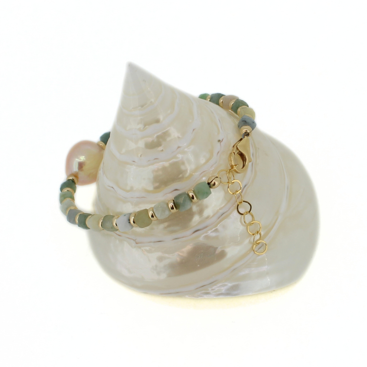 A beaded bracelet with green beads, a pale pink pearl bead, and gold spacer rings and clasp, displayed on a white seashell against a white background.