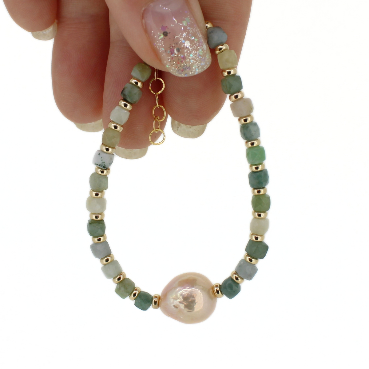 A beaded bracelet with green stones separated by gold spacer beads, featuring a large iridescent pearl at the bottom and held by a hand against a white background.