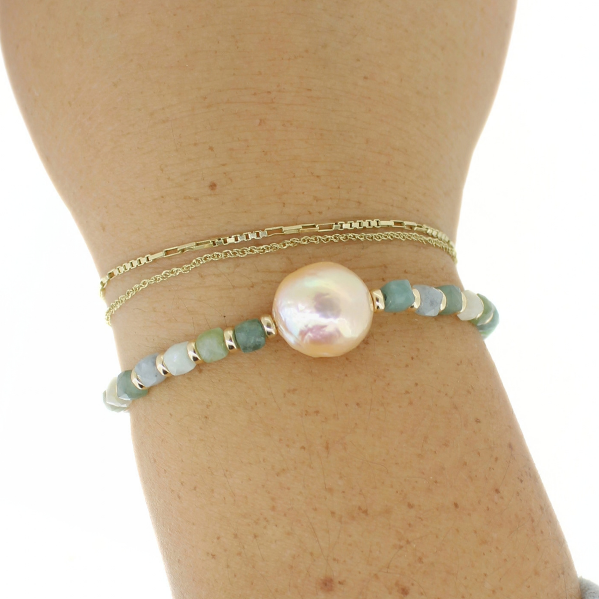 a wrist wearing delicate gold chain bracelets along with a green and gold beaded bracelet with a large white pearl at the center.