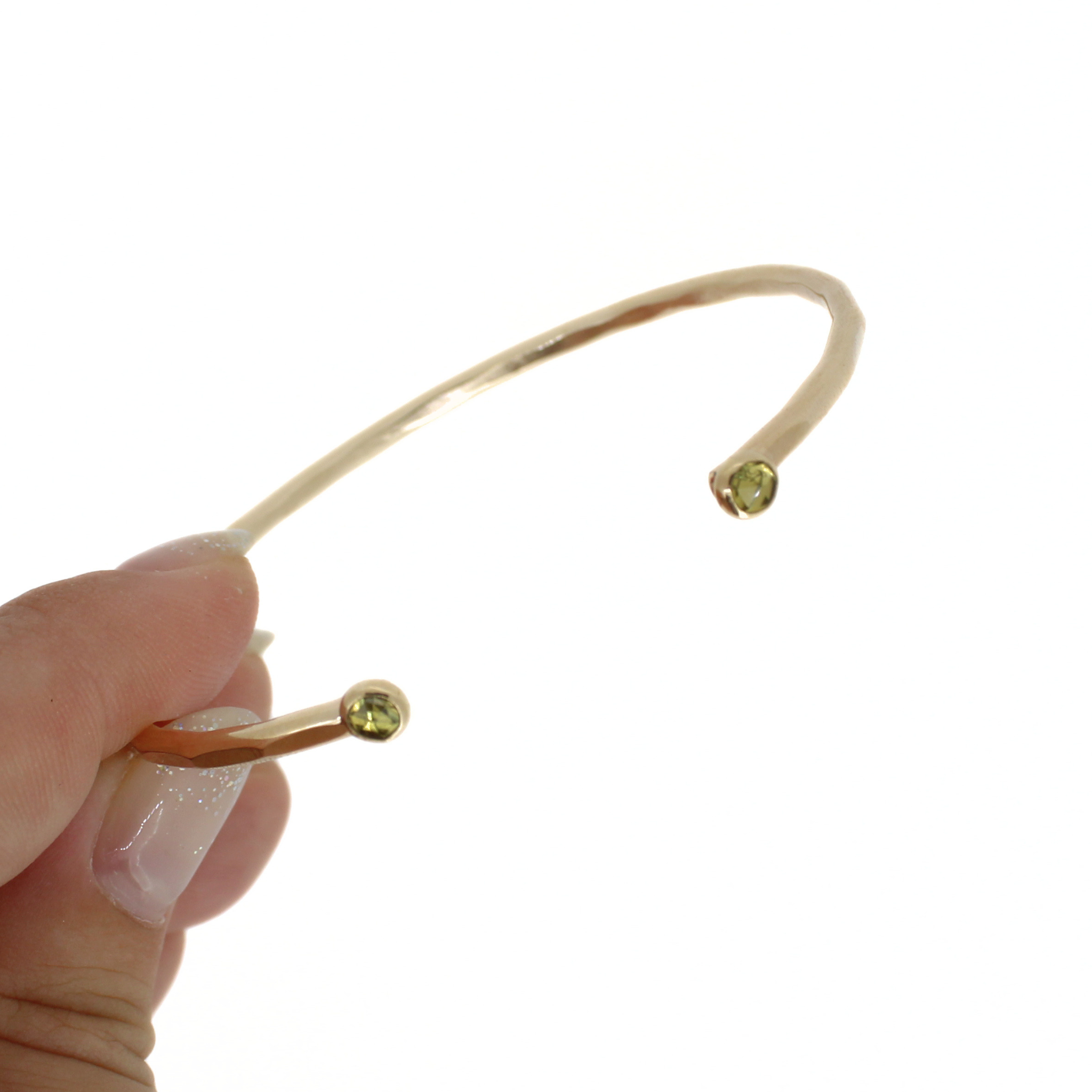A hand holding a thin open cuff bracelet made of yellow gold with two small faceted green peridot gemstone accents, shown against a plain white background.