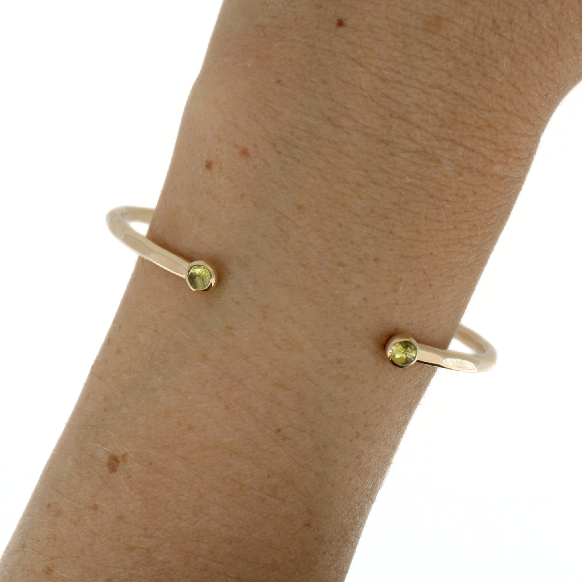 A close-up of a person's wrist wearing a gold cuff bracelet with small round green peridot gemstones set at the ends, against a plain white background.
