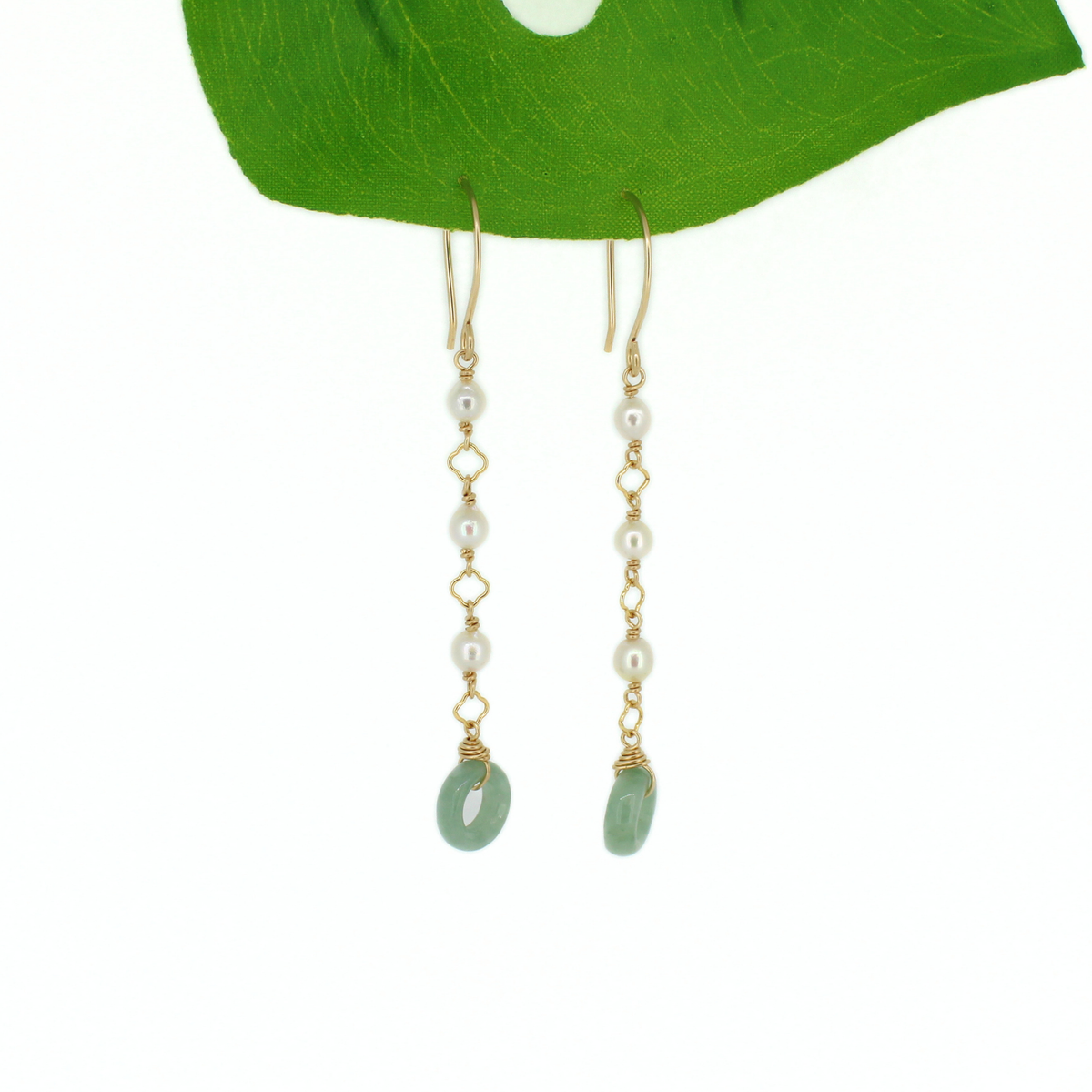A pair of gold drop earrings with white pearl beads and light green jade donuts, hanging against a white background beneath a green leaf.