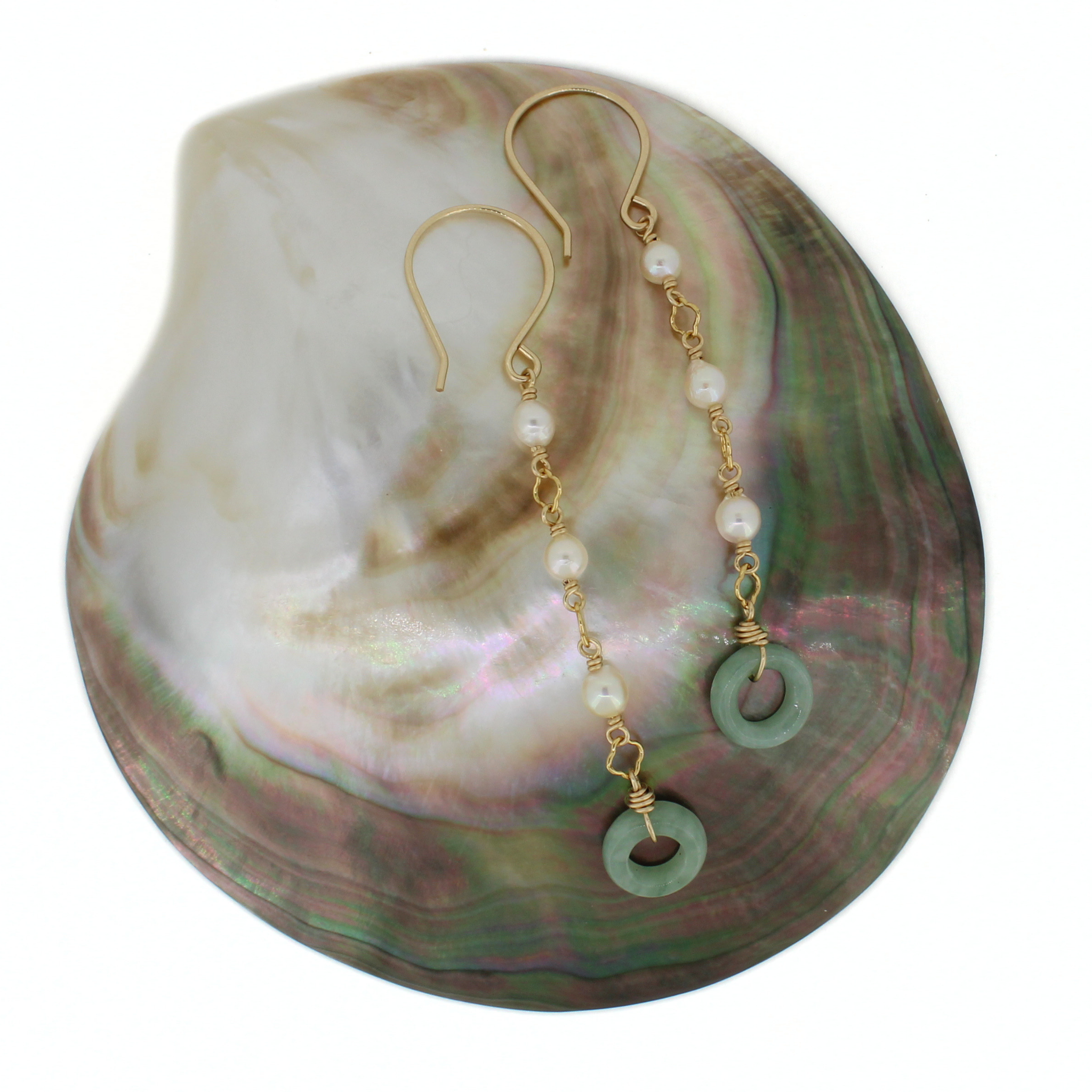 A pair of gold-tone dangle earrings with white pearl beads and green circular ring charms, displayed on a large iridescent abalone shell against a white background.