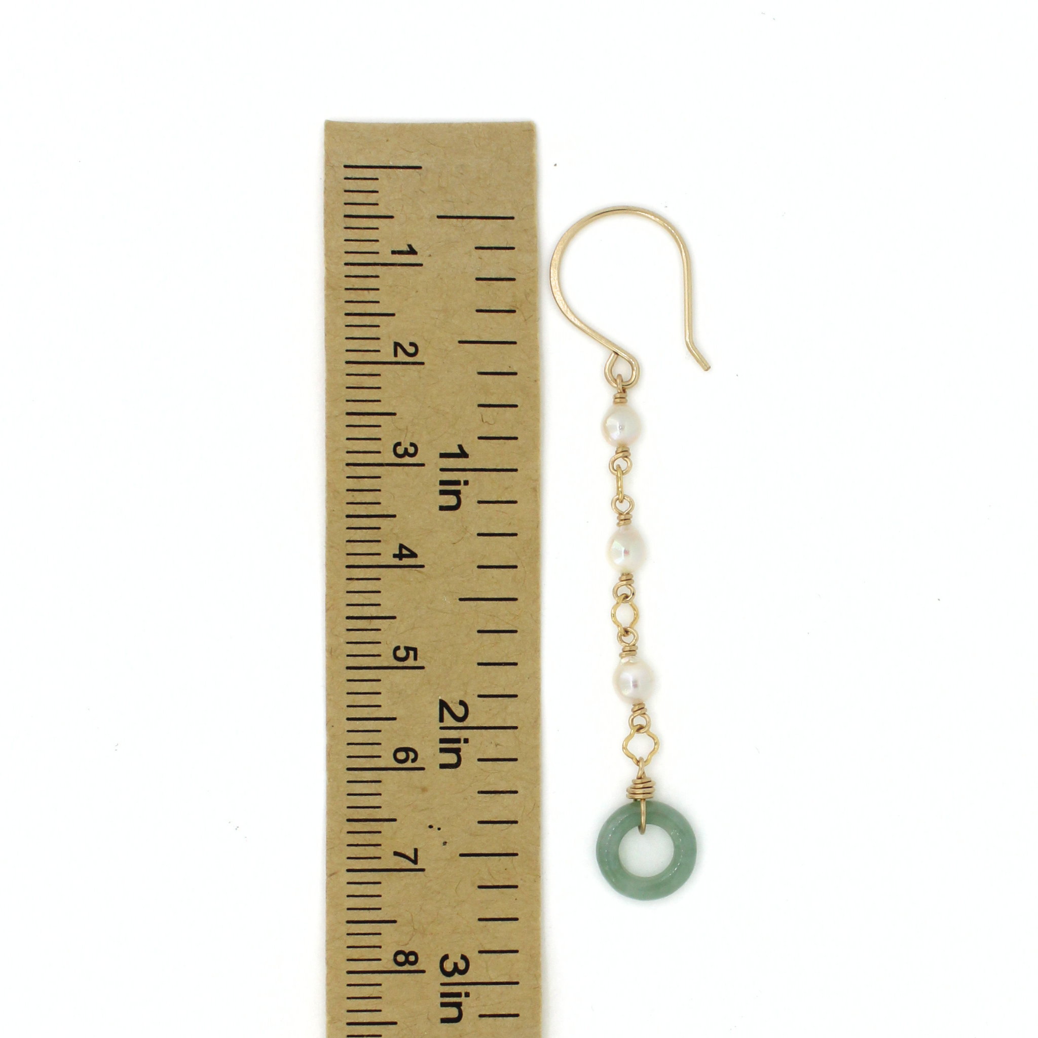 A single dangling earring shown next to a small ruler for scale on a white background. The earring has a gold fishhook ear wire, small white pearl beads, gold chain links, and a round light green jade donut at the end.