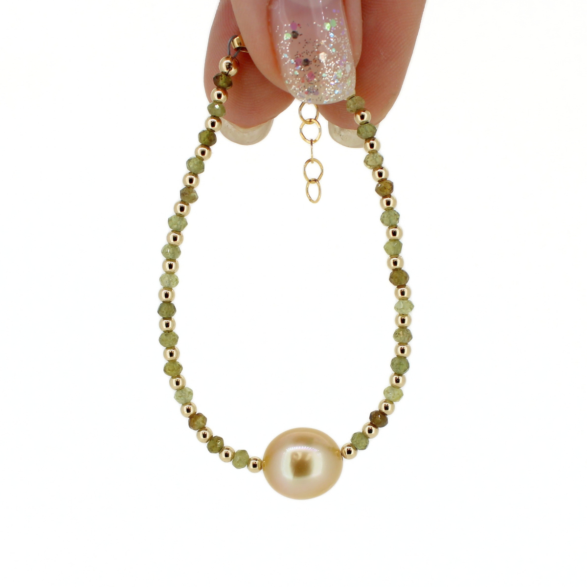 A delicate beaded bracelet with a large pearl at the center, held by a hand with glittery nail polish against a solid white background.