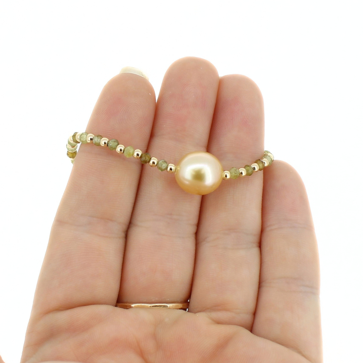 A delicate gold beaded bracelet with a central round pearl, shown resting on a person's hand against a plain white background.