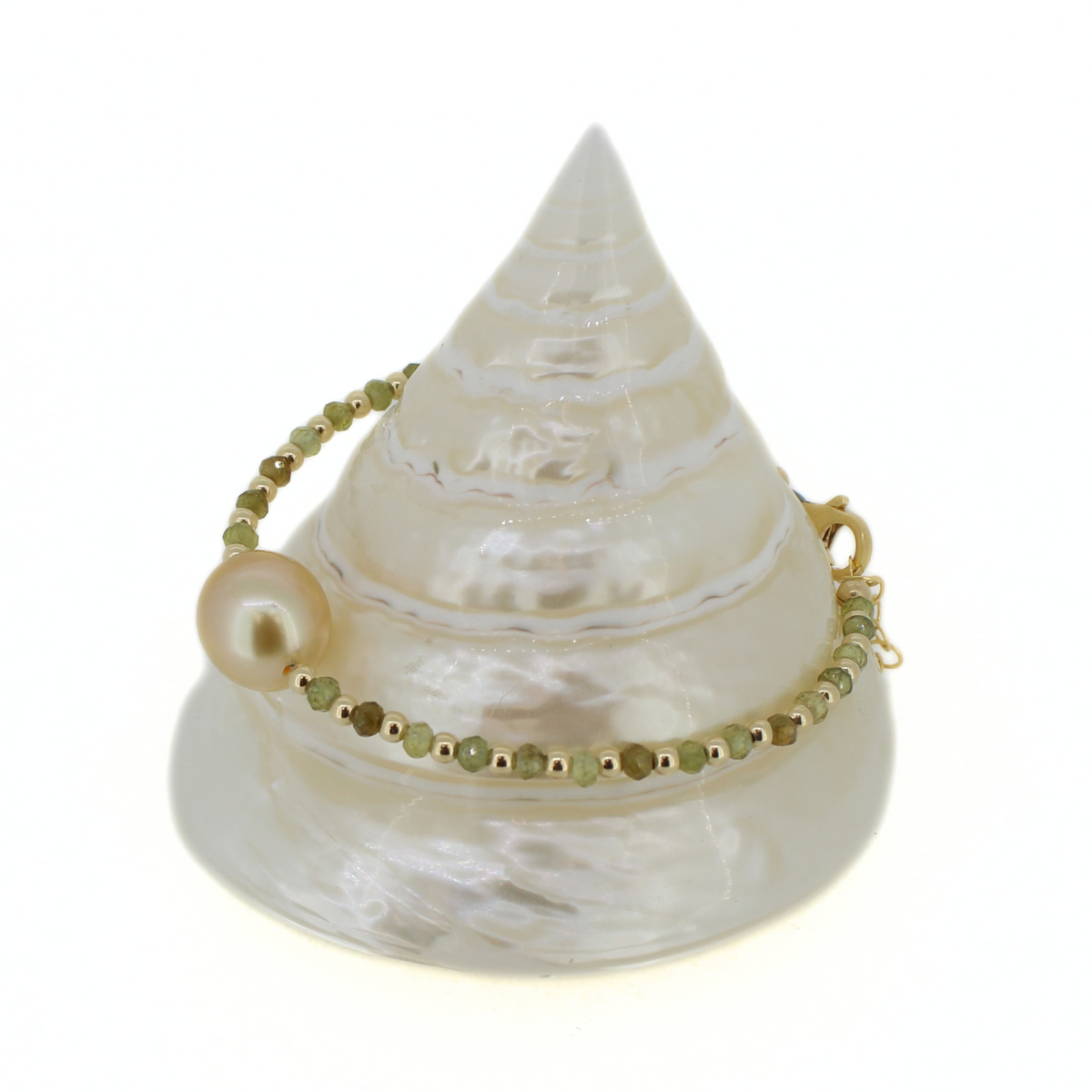 A delicate bracelet with a large pearl bead, small green faceted beads, and gold spacer beads, displayed on a white seashell stand against a white background.