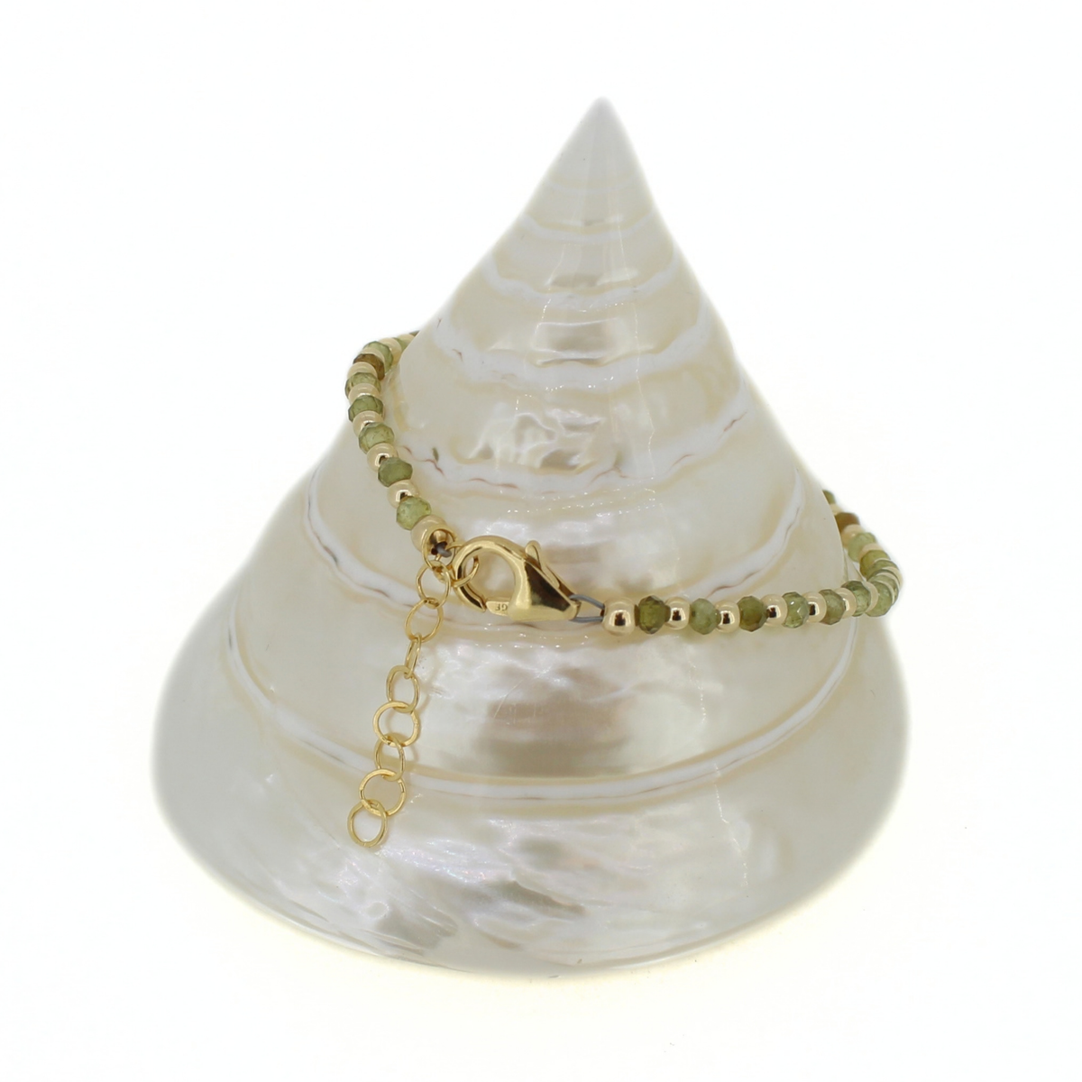 A delicate beaded bracelet with olive green beads and gold metal accents, with a lobster claw clasp and 1" extender. Displayed on a white seashell jewelry stand against a white background.