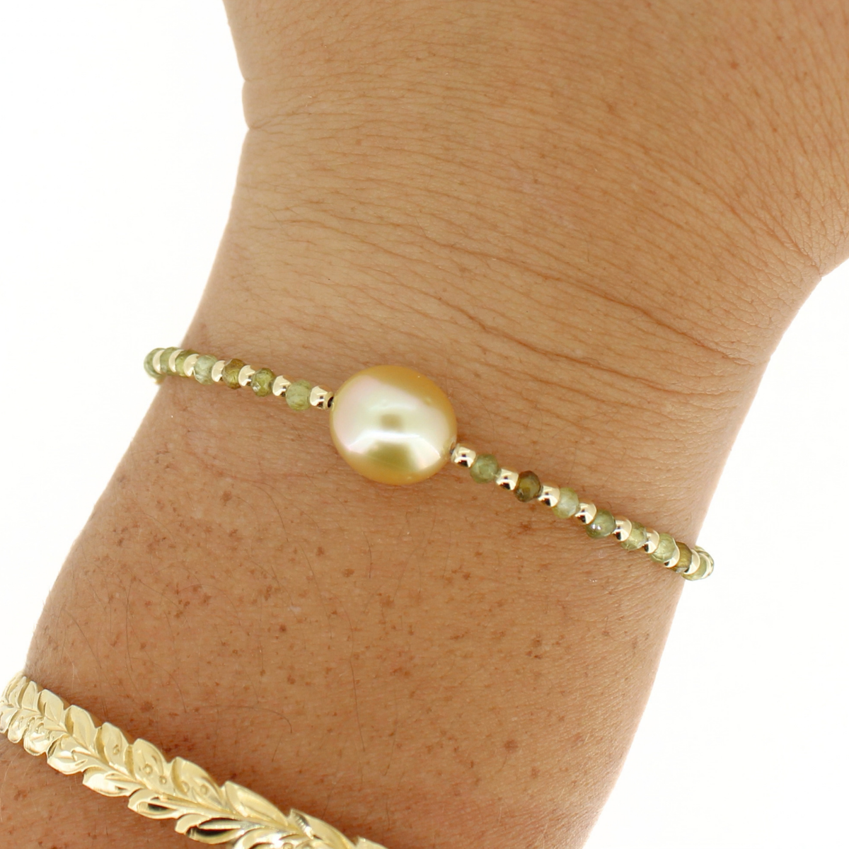 A gold beaded bracelet with a central golden south sea pearl, shown worn on a person's wrist alongside another gold bracelet.