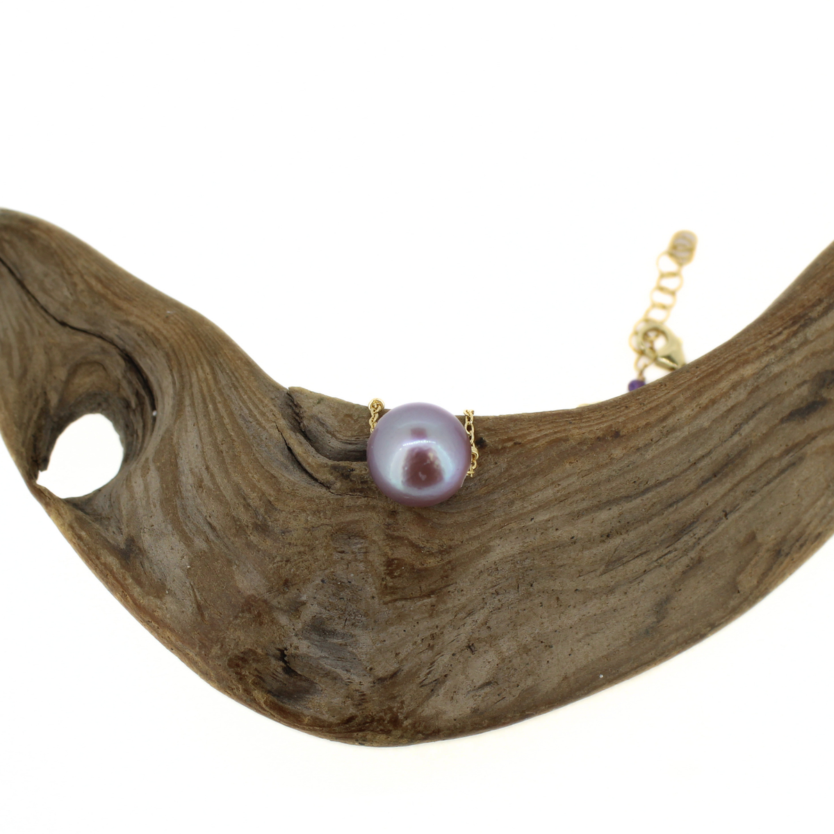 A gold necklace with a single round purple freshwater pearl, displayed on a curved piece of natural driftwood against a white background.